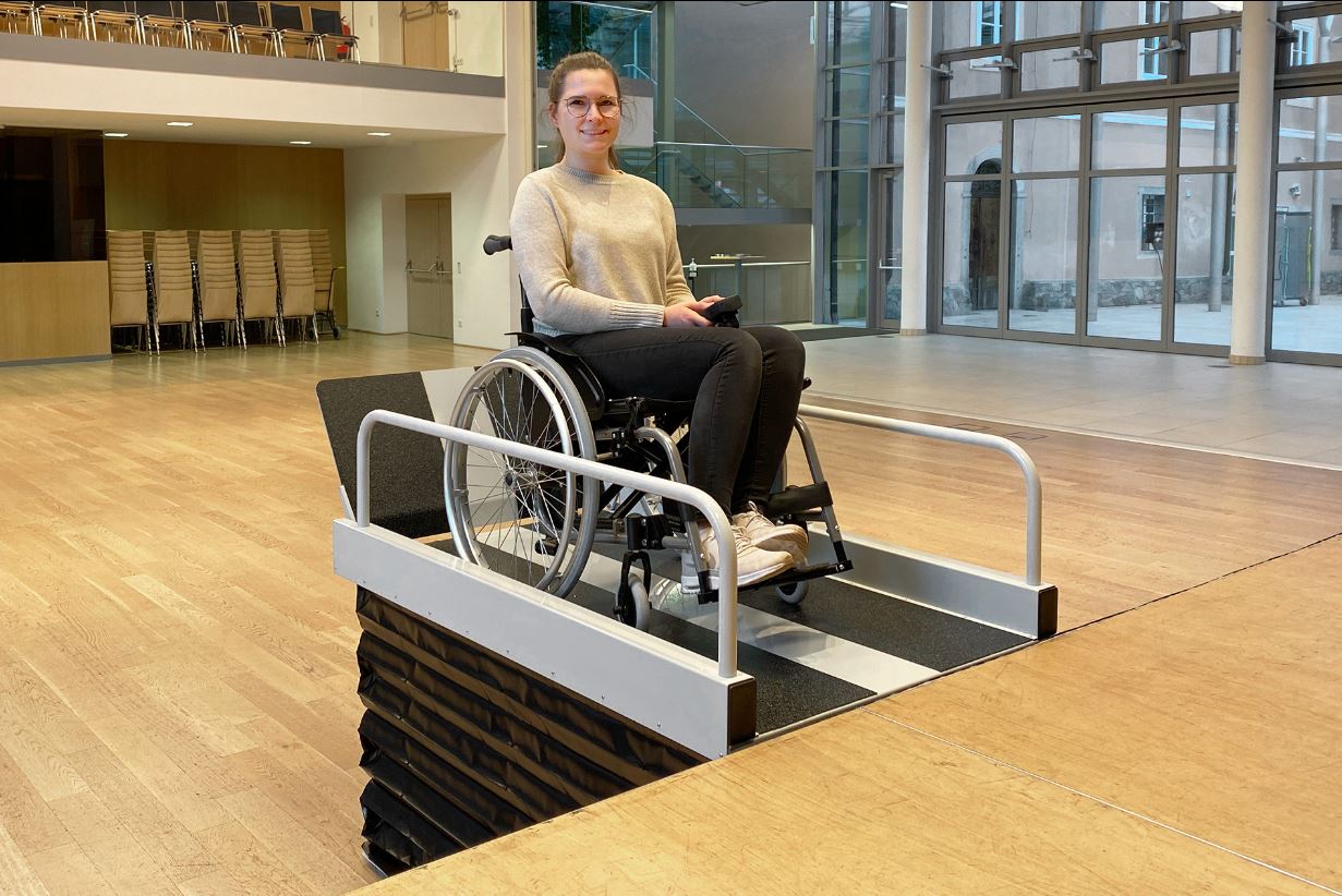 Liftboy in einem Saal. Eine Rollstuhlfahrerin ist auf Höhe der Bühne