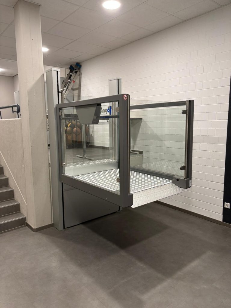 Plattformlift neben einer Treppe in einem Innenraum, mit Glaswänden und Metallboden.