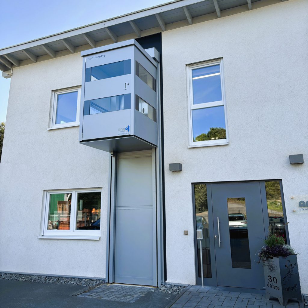 Senkrechtlift QuattroPorte vor einem Bürogebäude mit Eingangsbereich. Der Lift ist außen an der Fassade installiert und ermöglicht den stufenlosen Zugang zu mehreren Etagen.