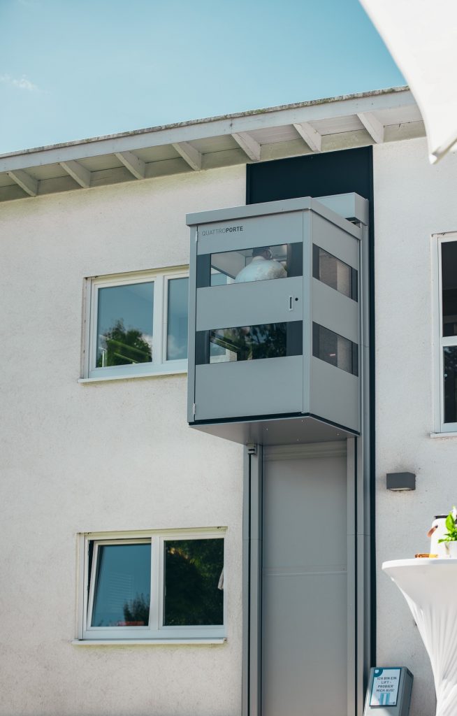 Moderner Senkrechtlift QuattroPorte an einer weißen Hausfassade, der bis ins Obergeschoss fährt. Der Aufzug ist teilweise verglast und außen am Gebäude angebracht.