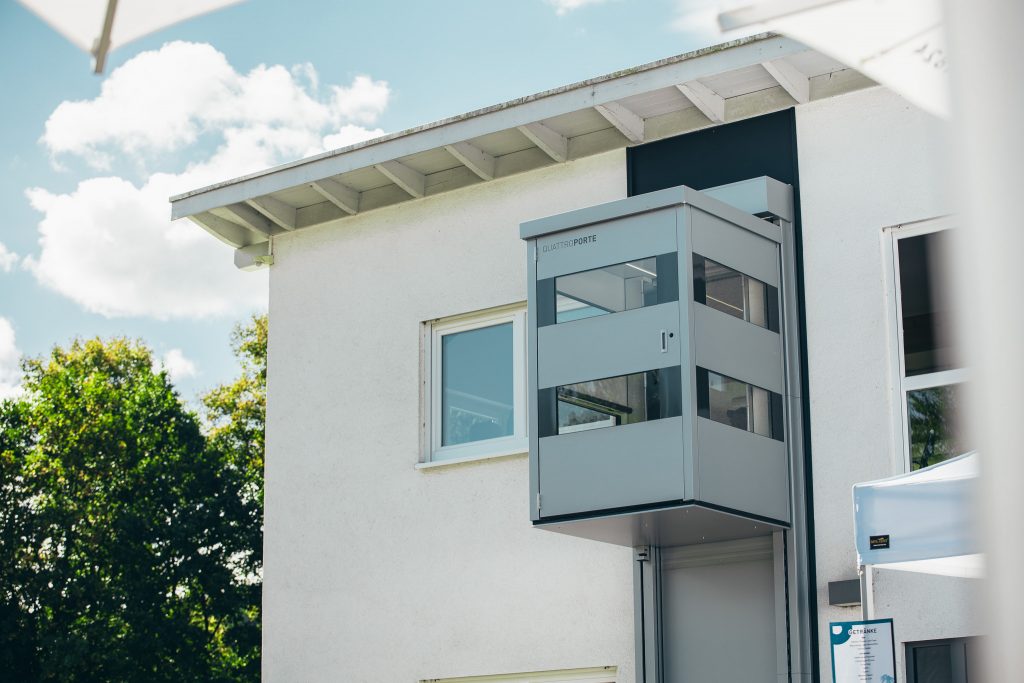 Außenansicht eines QuattroPorte Senkrechtlifts bei sonnigem Wetter. Der Lift verbindet Erdgeschoss und Obergeschoss eines Wohnhauses und bietet eine barrierefreie Lösung.
