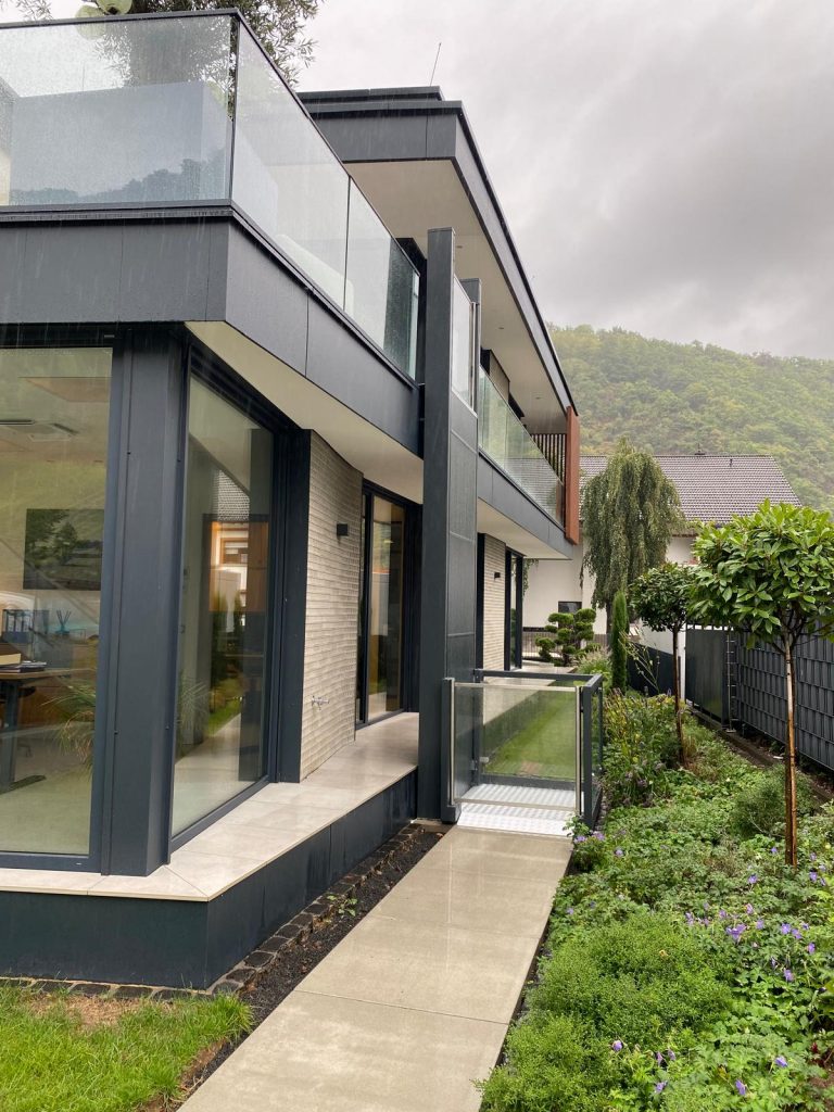 Plattformlift an der Außenseite eines modernen Hauses, verbindet den Gartenweg mit dem Balkon im Obergeschoss.
