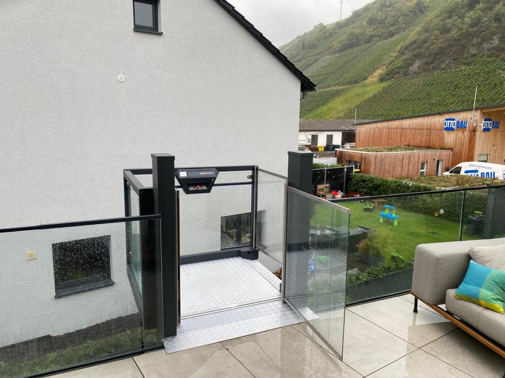 Plattformlift auf der Terrasse im oberen Stockwerk mit Glasgeländer, Blick auf Nachbargebäude und Weinberge im Hintergrund.