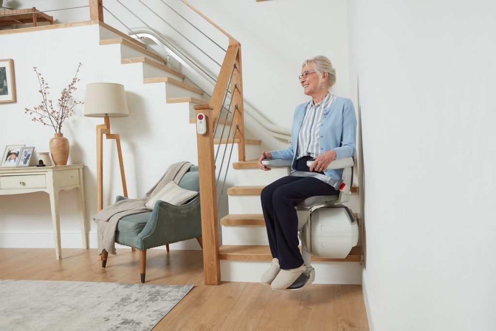 Eine ältere Frau, die zur Seite lächelt während Sie auf einem Ultimate Treppenlift unten an der Treppe sitzt.