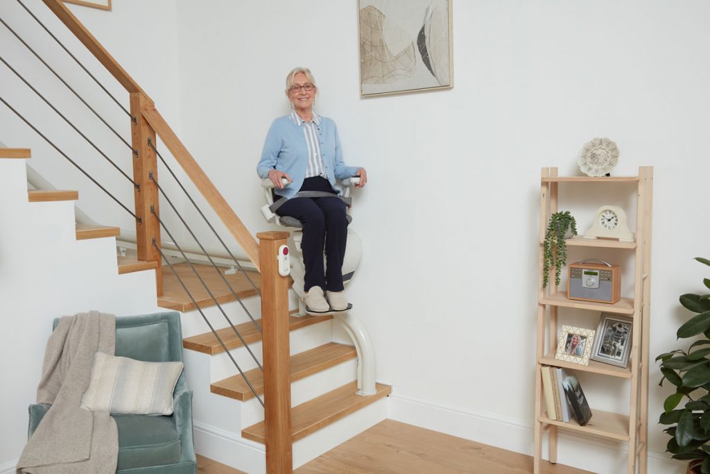 Eine ältere Frau, die in die Kamera lächelt während Sie mit einem Ultimate Treppenlift die Treppe runter fährt.