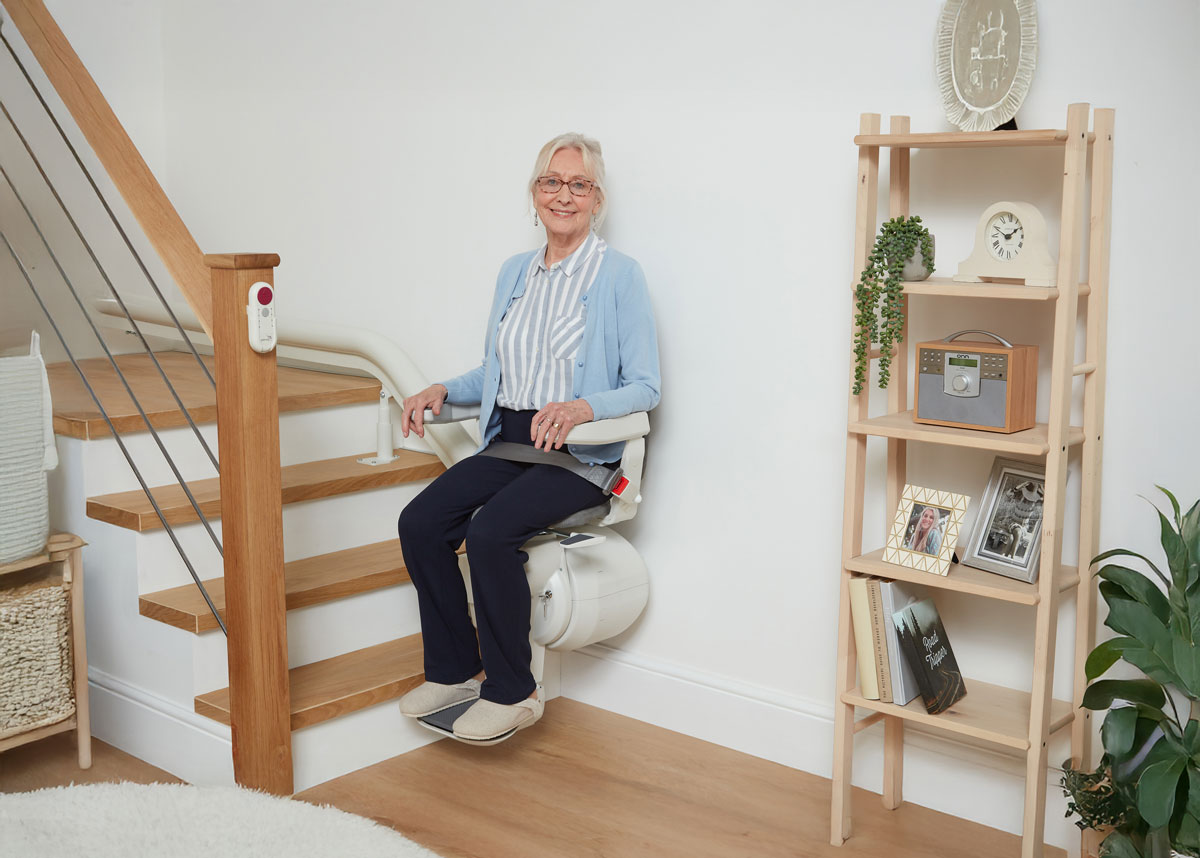Ältere Frau sitzt lächelnd auf einem Treppenlift in einem modernen Wohnraum mit Holztreppe und Regal im Hintergrund.
