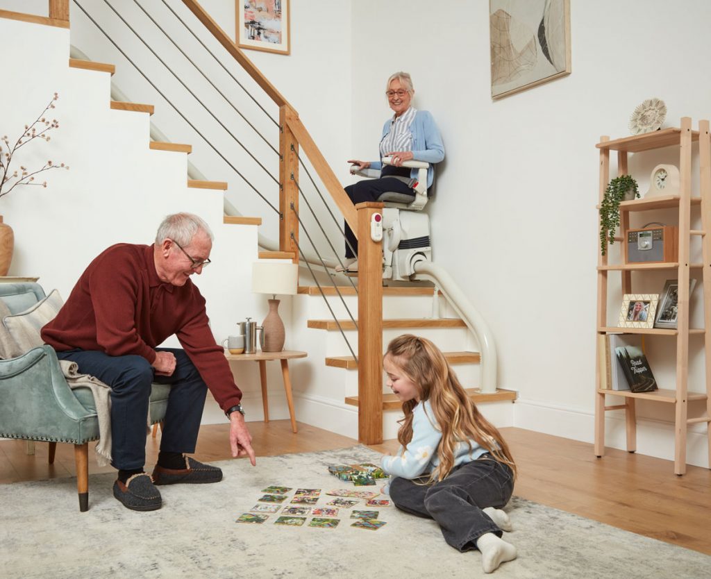 Großvater spielt mit seinem Enkelkind auf dem Boden, während die Großmutter mit dem Ultimate Treppenlift die Treppe hochfährt.