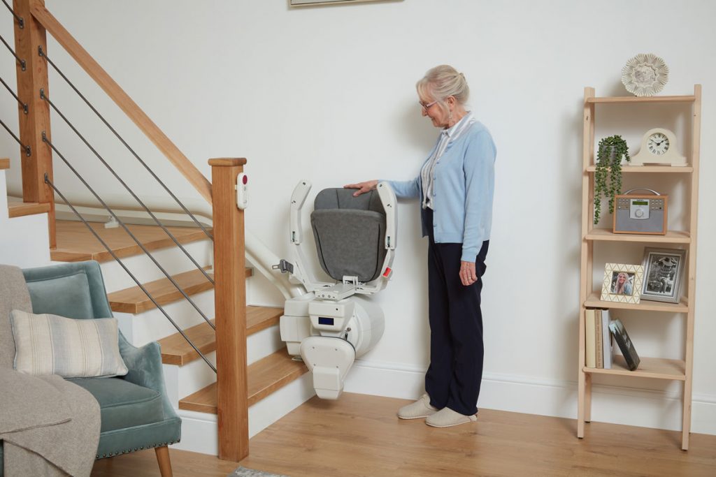 Eine ältere Damen, die ihre Hand auf die Lehne eines grauen Ultimate Treppenlifts legt.