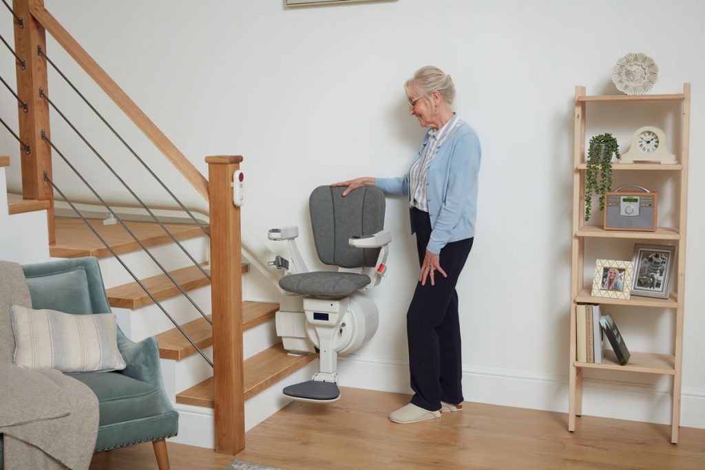 Eine ältere Damen, die ihre Hand auf die Lehne eines grauen Ultimate Treppenlifts legt.